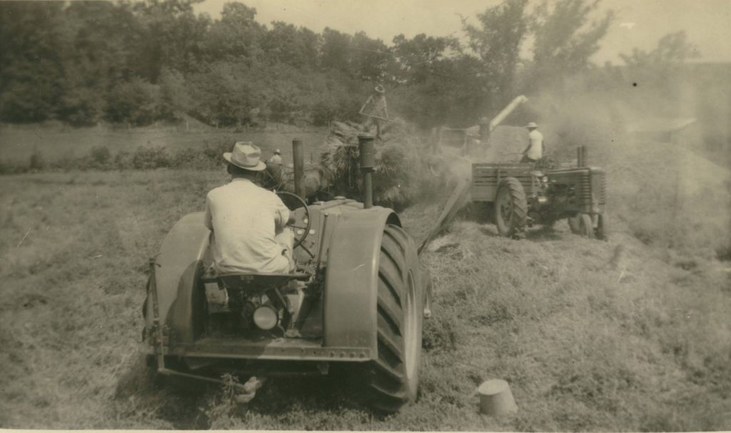 Black and white photograph of a man driving a tractor in a field with other men threshing in the background