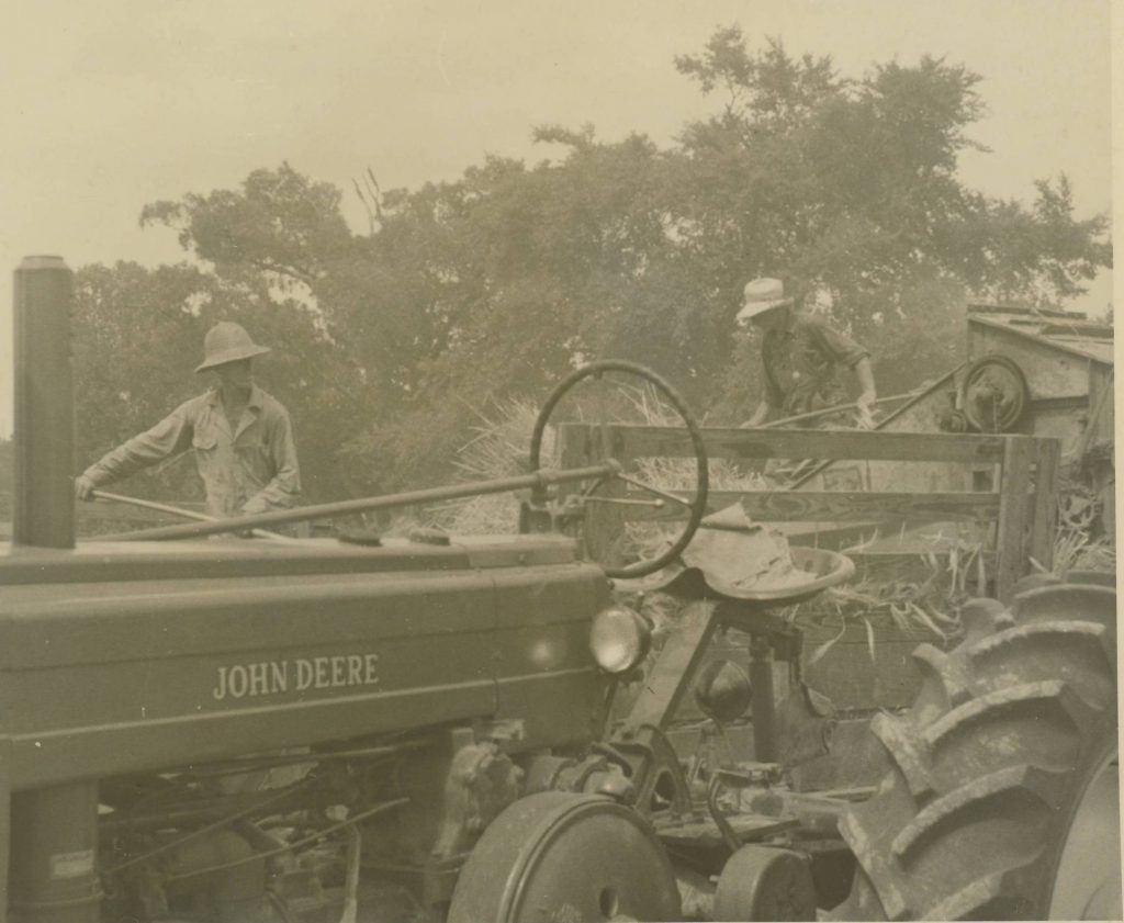 Black and white photo of two men threshing in a field with a John Deere tractor in the foreground